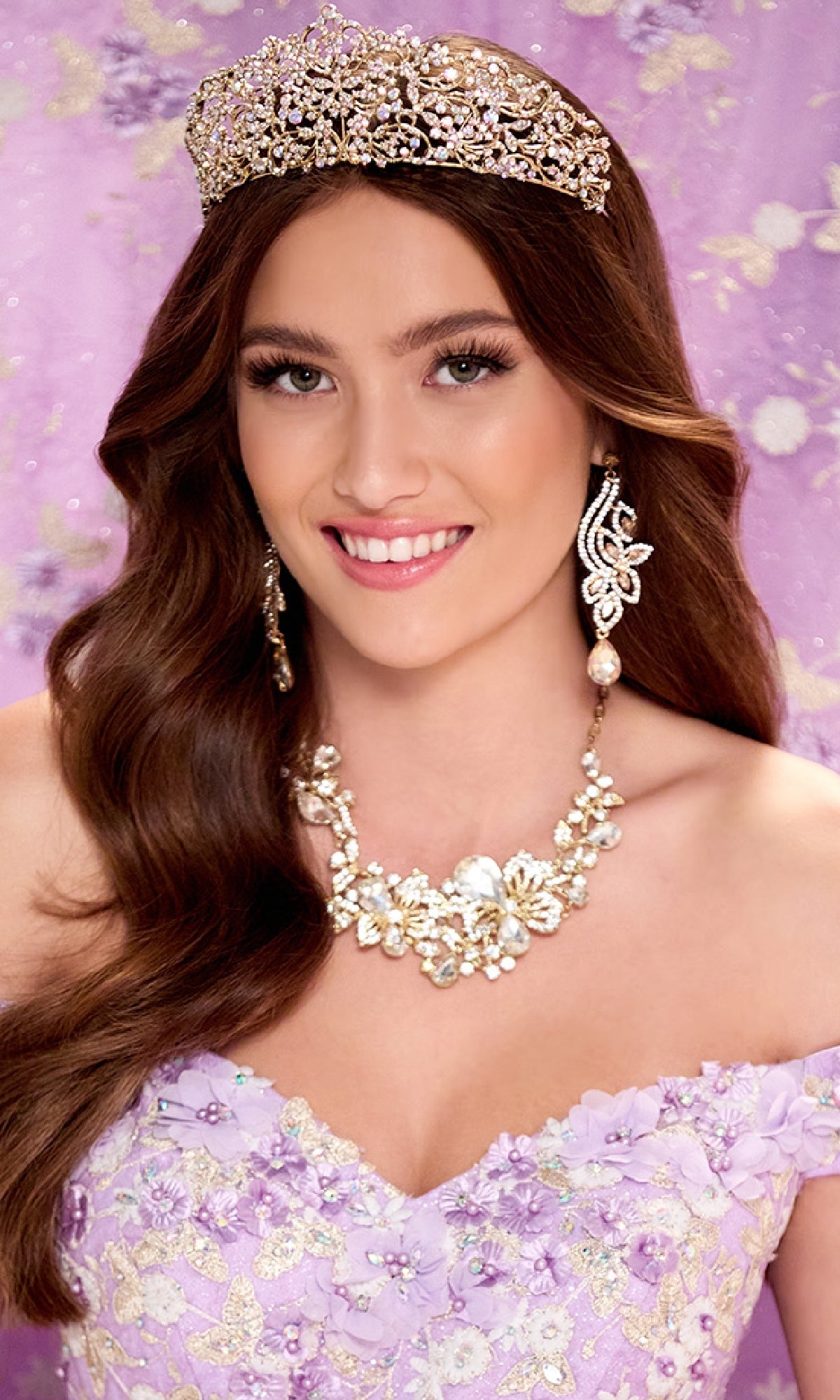 A woman smiles in the Quinceanera Dress PR30084 By Princesa—a jeweled crown, ornate earrings, and matching necklace accent her lavender, flower-embellished gown with a corset back, set against a purple floral backdrop.